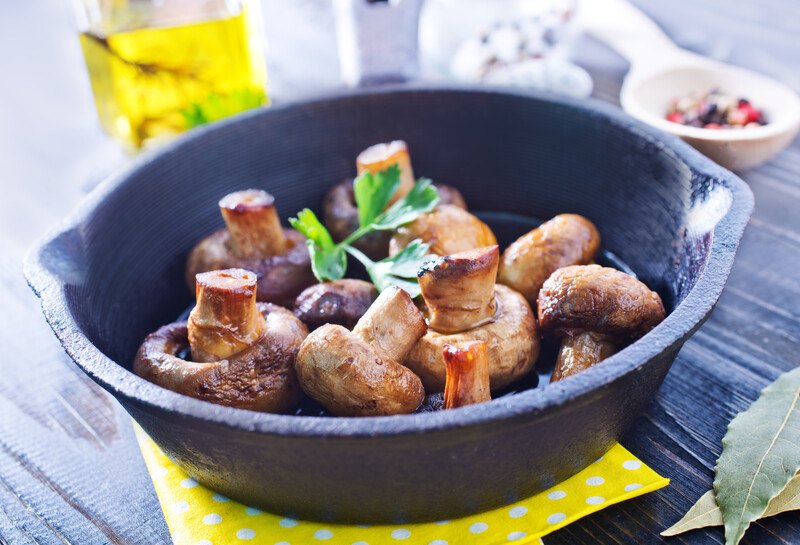 on a wooden table is a skillet with roasted mushroom, underneath is a yellow table napkin with laurel leaves beside it, at the back is a bottle of olive oil and a wooden spoon full of peppercorns at the back