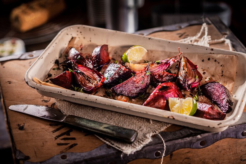 on a wooden board with metal linings, is a rectangular brown ceramic dish with roasted red onions with sliced limes, underneath is a piece of burlap and a knife beside it