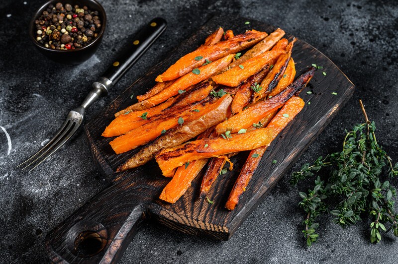 on a dark wooden chopping board are roasted sweet potato sticks, beside it is a sprigs of herbs, a fork, and a tiny bowl of peppercorns