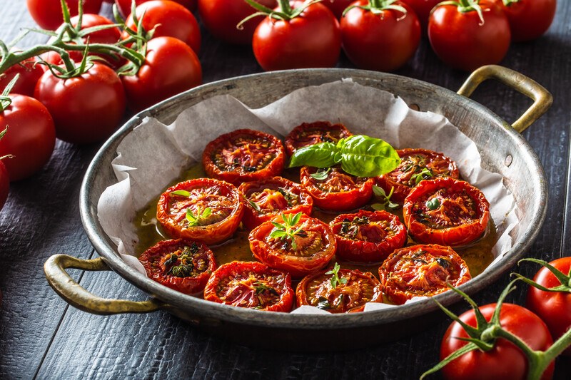 roasted sliced tomatoes in a shallow metal pan with baking paper with fresh tomatoes around it