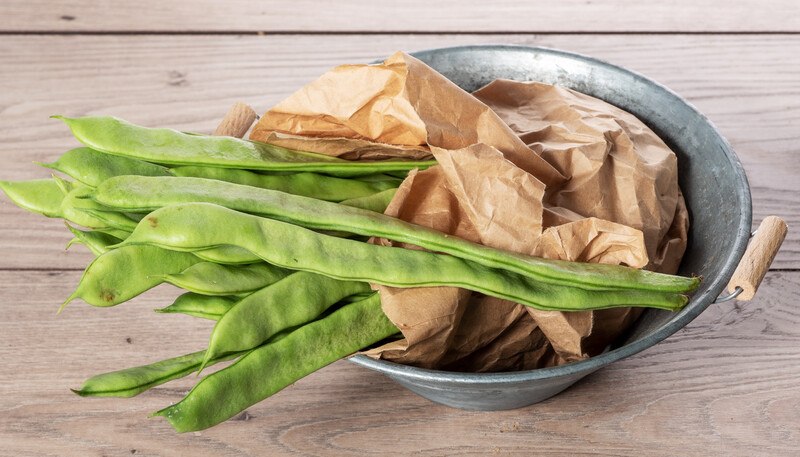 on a wooden surface is a tiny metal wok with fresh Romano beans in it partially wrapped in brown paper