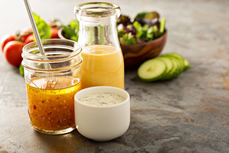 closeup image of different containers with different salad dressings, with a bowl of green salad, sliced cucumber, and cherry tomatoes at the back 