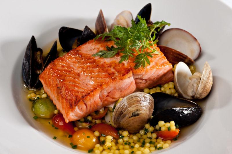 salmon fillet on top of a bed of couscous, with clams peppers and tomatoes on a white bowl.