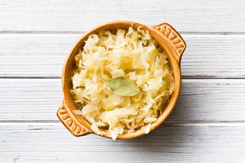 A wooden bowl filled with sauerkraut and a bay leaf rests on a light wooden surface.