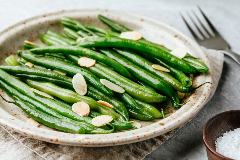 on a concrete surface is a rustic looking ceramic plate with sauted green beans with slivered almonds on top resting on a tan-colored table napkin, beside it is a silver fork and a small dish of rock salt