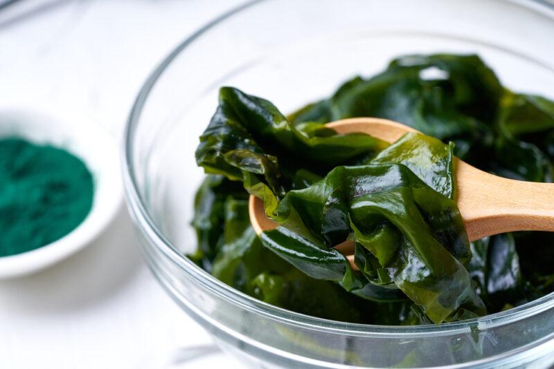 a closeup image of a clear glass bowl with green seaweed and a wooden spoon shoved in it, beside it is a little dish with powdered seaweed