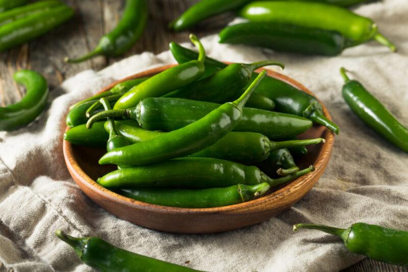 a wooden brown dish full of green serrano peppers resting on a piece of beige kitchen towel with loose serrano peppers around it