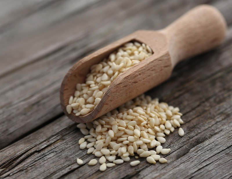 closeup image of a wooden scoop full of sesame seeds with a small heap of sesame seeds beside it, resting on an aged wooden surface