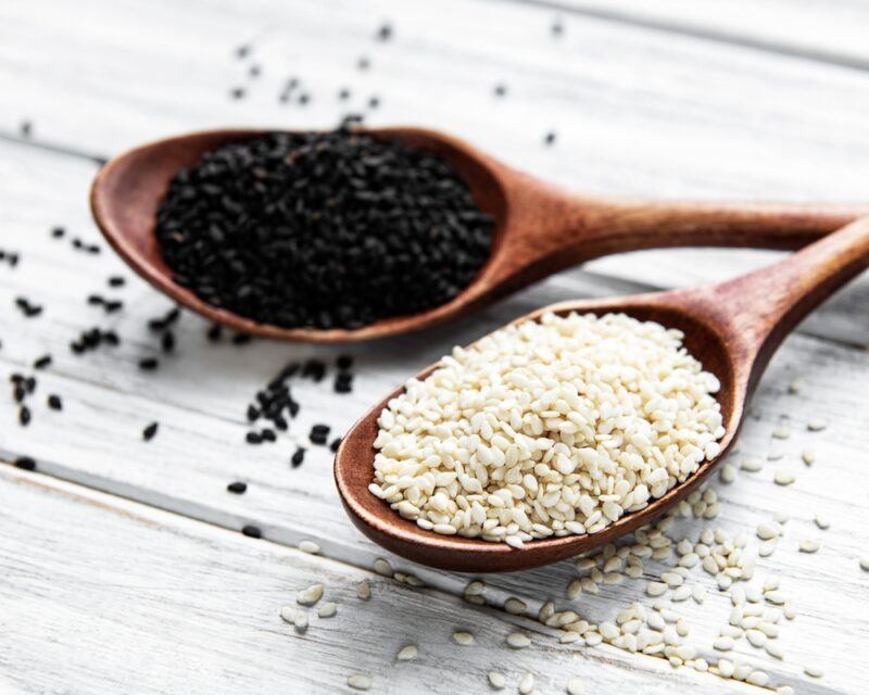 on a white wooden surface is a couple of dark brown wooden spoon with white and black sesame seeds, around it are loose sesame seeds