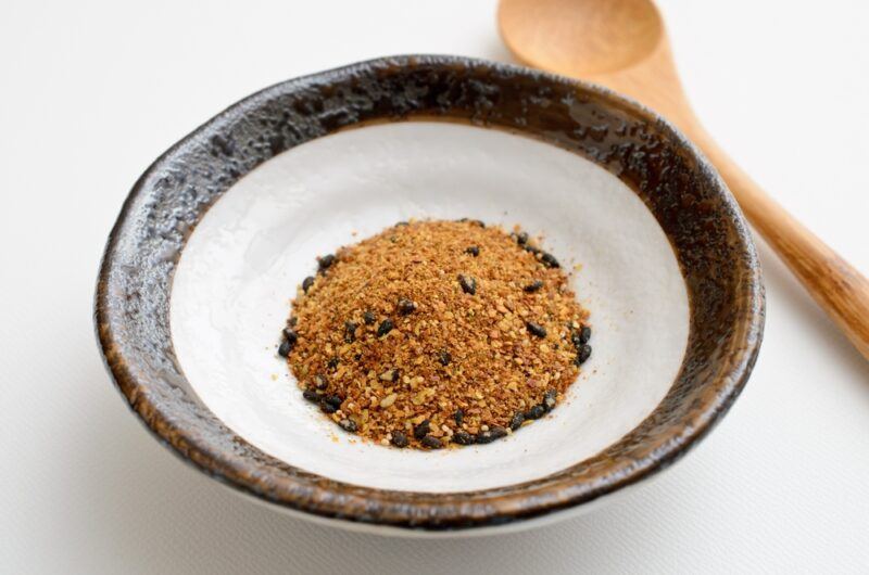 on a white surface is a white and dark brown ceramic bowl with shichimi togarashi, with a wooden spoon beside it