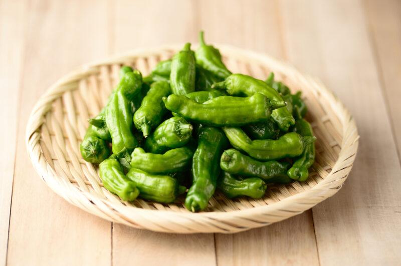 on a wooden surface is a light brown weaved shallow tray full of shishito peppers