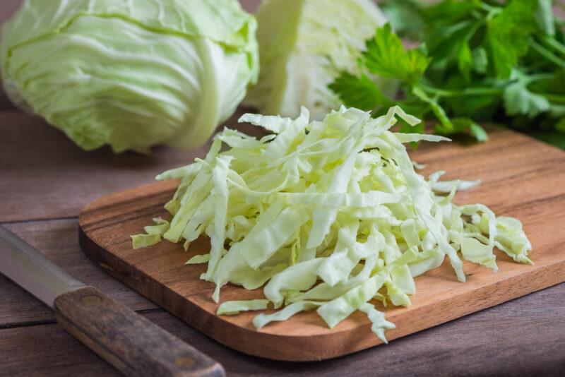 on a wooden surface is a wooden chopping board with shredded cabbage, beside it is a knife, at the back are whole cabbage head and a bunch of parsley