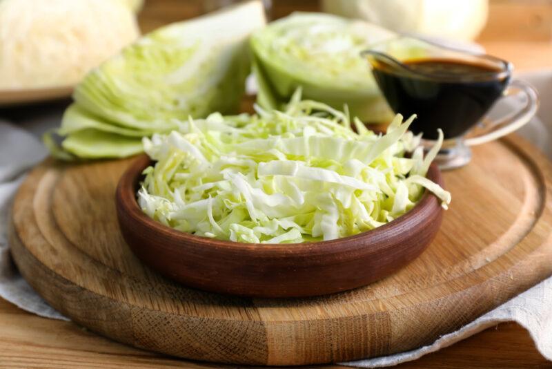 on a thick wooden chopping board is a wooden bowl of shredded cabbage with quartered cabbage and clear glass container of balsamic sauce at the back