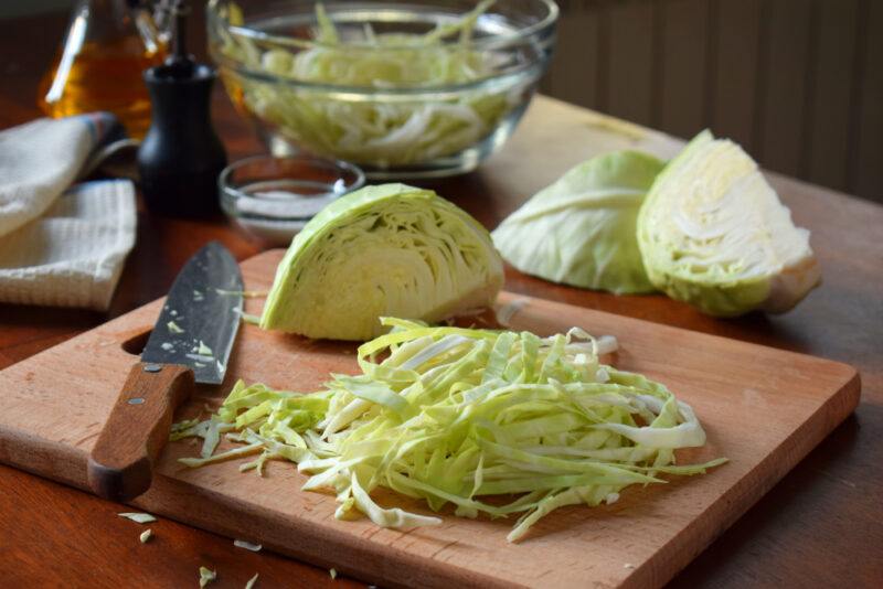 cut and shredded cabbage on a wooden chopping board with a knife beside it and a bowl of shredded cabbage at the back