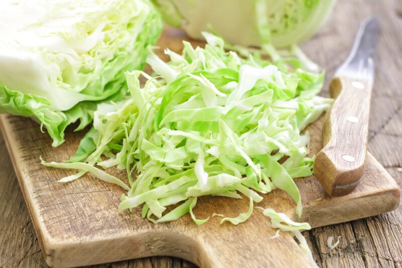 on a wooden surface is a closeup image of wooden chopping board with a quartered cabbage, one partially shredded with a knife beside it