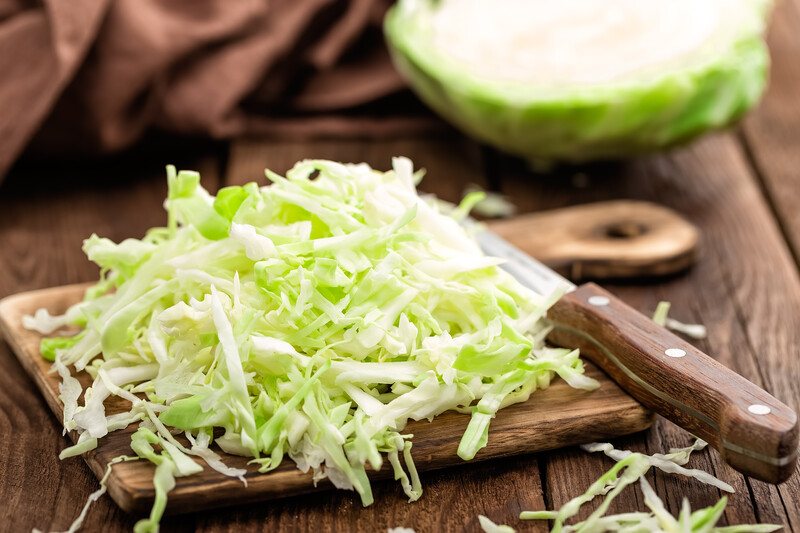 on a wooden surface is a closeup shot of shredded cabbage on a wooden chopping board with a knife beside it and halve cabbage head and brown table napkin at the back