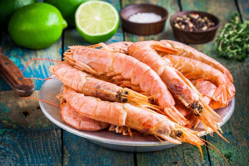 on a wooden surface is a white dish full of shrimp with lime, spices, and herbs at the back