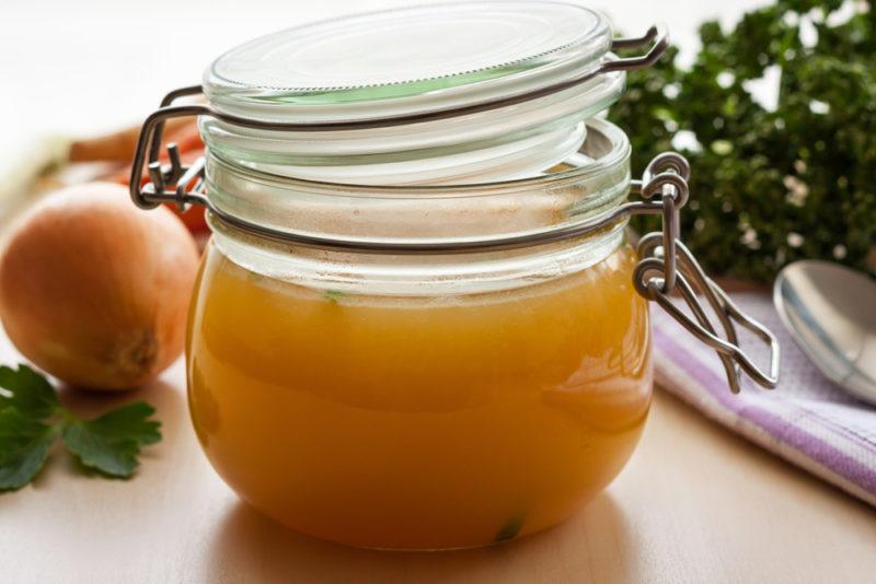 A jar of bone broth with an onion and herbs in the background