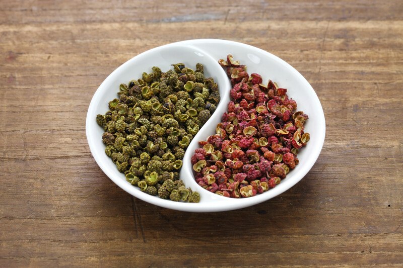 on a wooden surface is a white dish with half of it filled with green Sichuan peppercorns and the other half with red Sichuan peppercorns