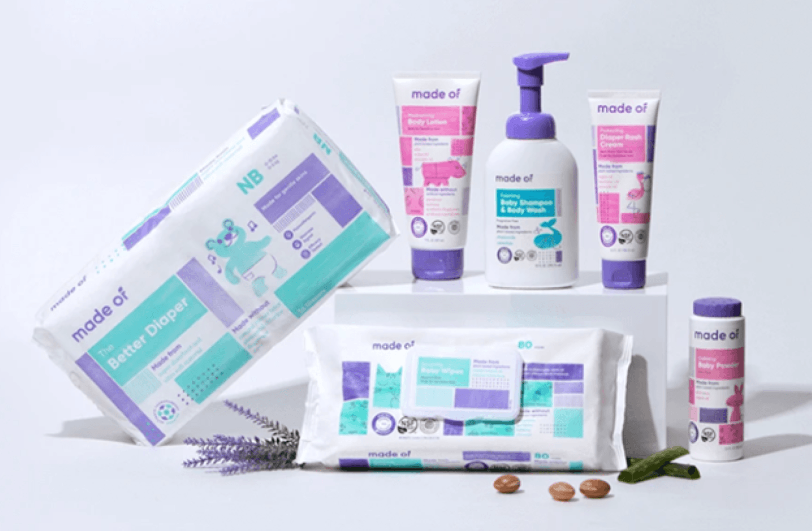 six different baby products diapers, wipes, lotion, shamoo, diaper rash cream, and diaper powder sutting on a white background with fresh lavender, shea cocoa beans, and pieces of aloe sitting amongst the products