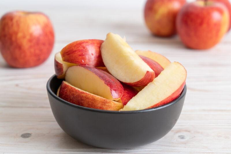 white wooden surface with a black bowl full of sliced red apples, at the back are a few whole red apples