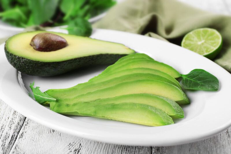 on a rustic looking white wooden surface is a white plate with sliced avocado and a halved avocado, at the back is a sprig of cilantro and a sliced lime