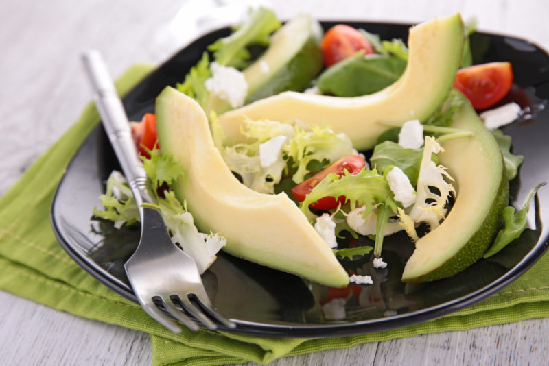 sliced avocado salad with lettuce and cheese | Food For Net a black plate filled with avocado salad composed of sliced fresh avocado, arugula, cherry tomatoes and feta cheese