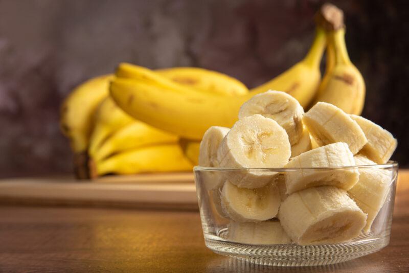 on a wooden surface is a clear glass dish with sliced bananas, at the back are fresh yellow bananas