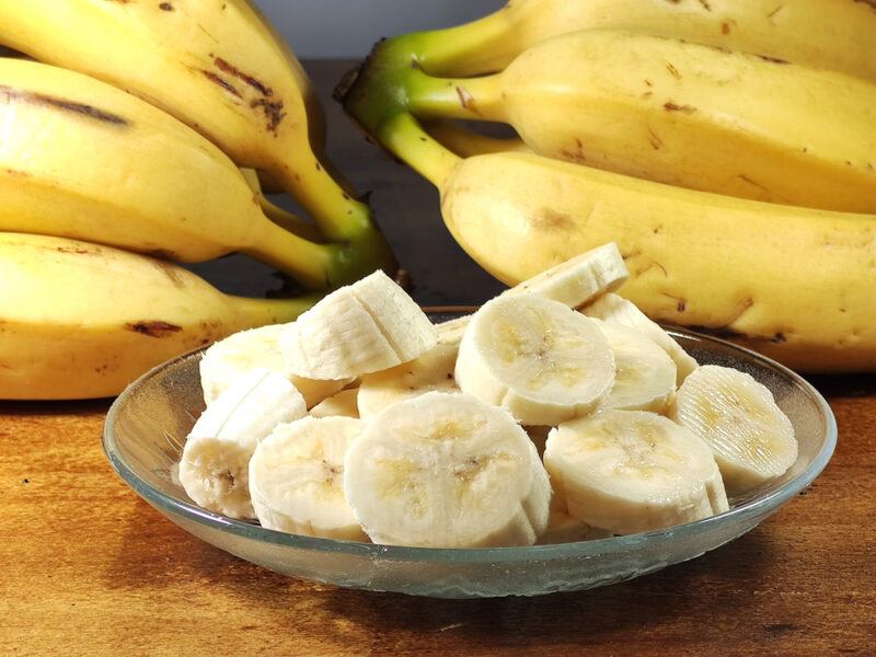 on a wooden surface is a clear glass saucer with sliced bananas, at the back is a couple of hands of bananas