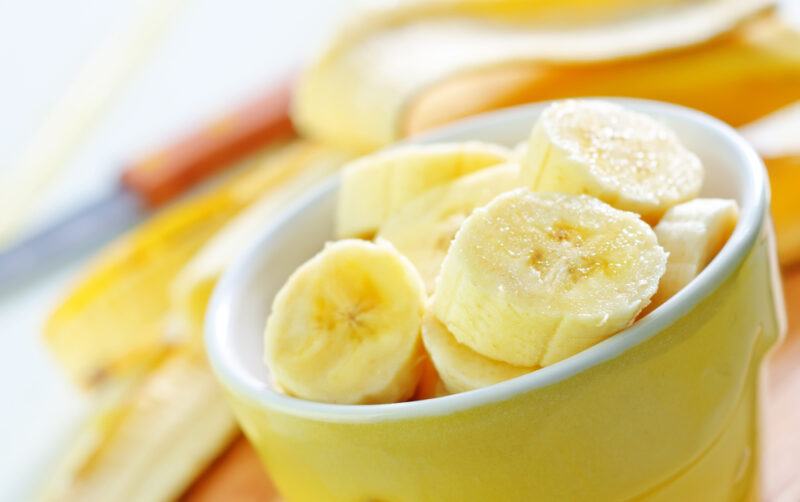 a yellow bowl with white rim full of sliced banana, at the back is a half-way peeled banana