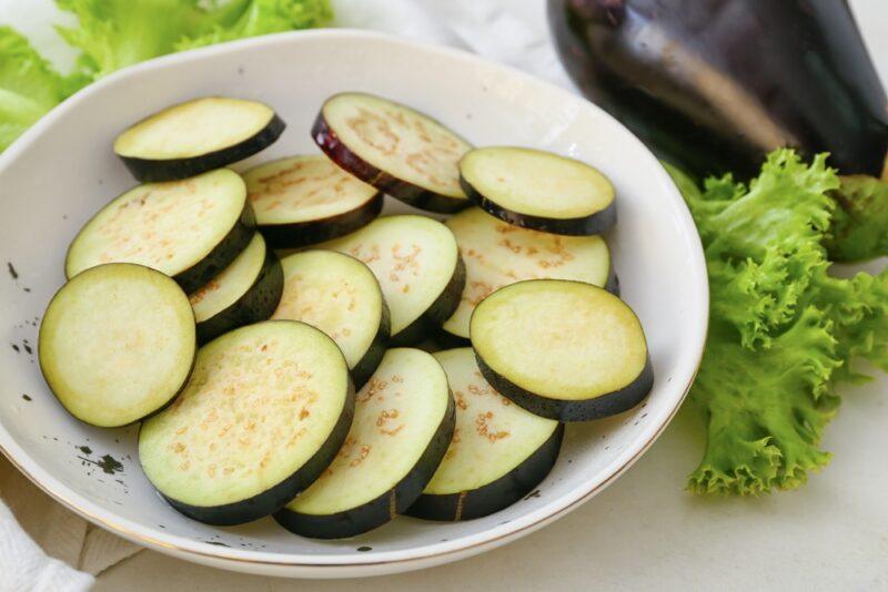 closeup image of a white bowl with sliced eggplant, with whole eggplants and a couple of lettuce leaves beside it