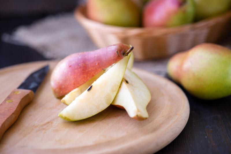 on a round wooden chopping board are sliced pears and a knife, at the back is a basket of whole pears with one loose pear