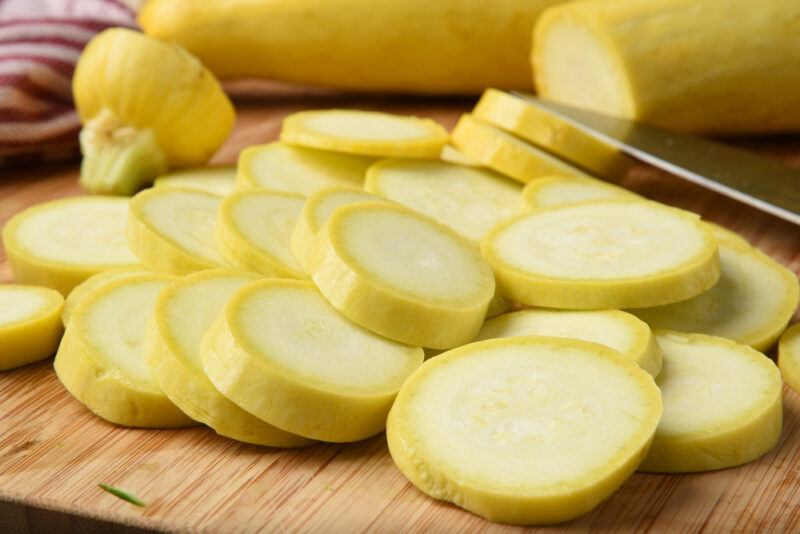 a closeup image of sliced summer squash on a chopping board, a couple of whole summer squash at the back