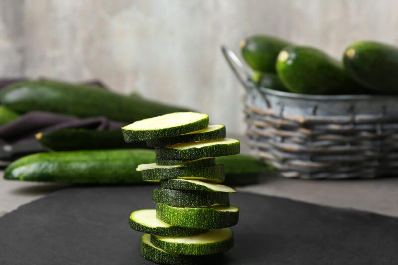 a pile of sliced zucchini on a black stone slab with basket full of zucchinis and loose whole zucchinis at the back