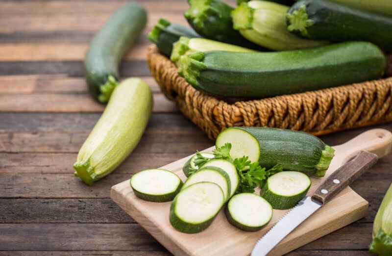 on a wooden surface is a wooden chopping board with sliced zucchini with a sprig of parsley and a knife, at the back are a couple of zucchinis beside a basket full of zucchinis