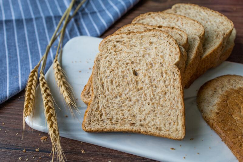 on a dark wooden surface are slices of whole wheat bread resting on a rectangular marble board with a single oat stalks and blue table napkin with white stripes 