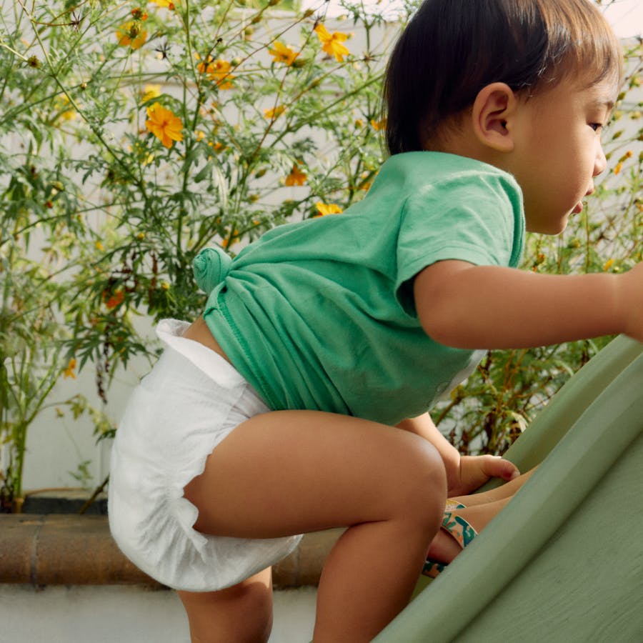 small child in a green shirt and white diaper climbing up a slide - with flowering plants in the background