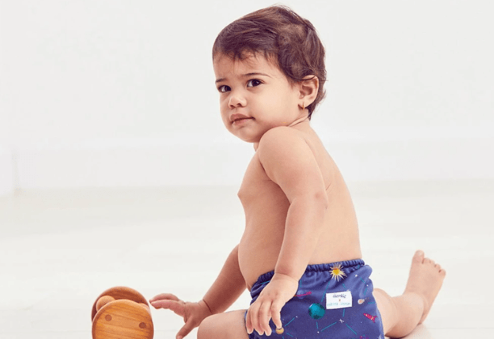 small child sitting in a cosmic designed diaper with a small wooden toy on a white background