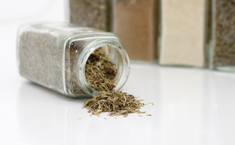 on a white surface is a topple jar of dried thyme with contents pouring out, at the back are lined up jars of different dried spices and herbs