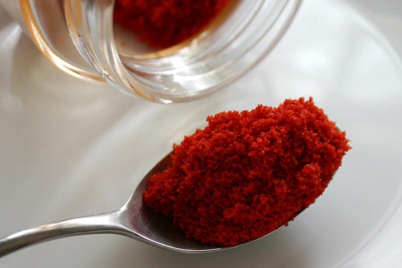 a closeup image of a silver spoon with smoked paprika powder, resting on a white plate with the jar of paprika powder closely beside the spoon