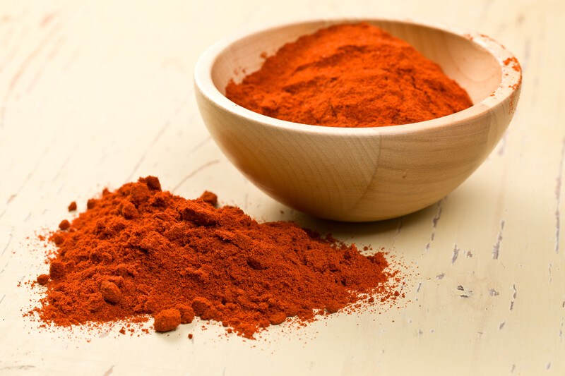 a light brown surface with a mound of smoked paprika with a wooden bowl full of smoked paprika beside it