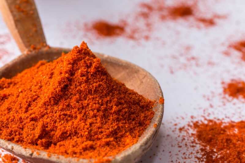 on a white surface is a closeup image of a wooden spoon full of smoked paprika with loose paprika powder around it