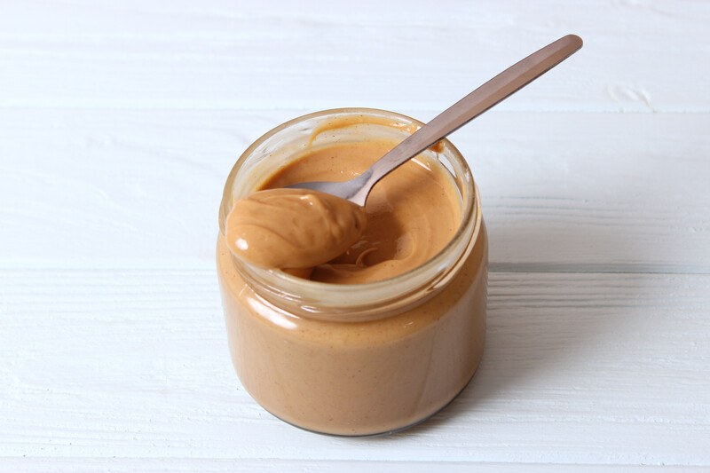 smooth peanut butter | Food For Net on a white wooden surface is a small jar of smooth peanut butter with a silver spoon on top also full of peanut butter