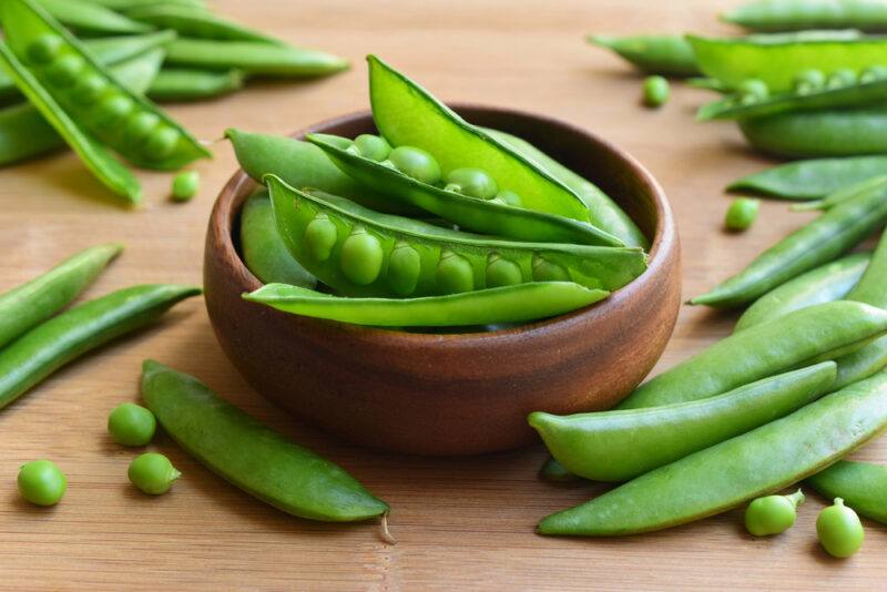 on a brown wooden bowl is a wooden bowl full of snap peas, with loose snap peas around it