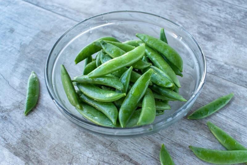 on an aged wooden surface is a clear glass bowl with fresh snap peas with loose snap peas around it