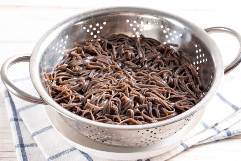 on a wooden surface is a white dish cloth with blue lines with a metal colander containing boiled soba resting on a white wide-mouthed bowl.