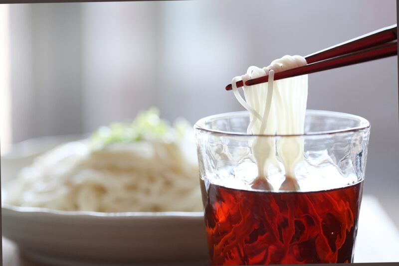 a closeup image of somen noodles in a white dish at the background and chopsticks with somen noodles getting dipped in tsuyu in the foreground