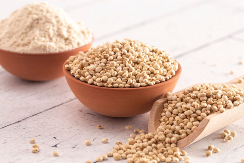 on a wooden surface is a couple of clay bowls, one with sorghum flour and the other with sorghum grains, beside it is a wooden scoop with overflowing sorghum grains