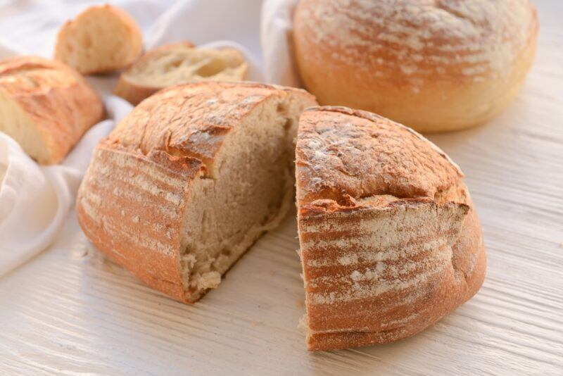 on a white wooden surface is a halve sourdough bread with a whole bread at the back and a few slices on a white table napkin