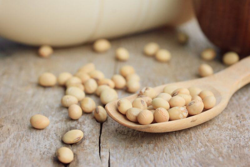 on a rustic wooden surface is a wooden ladle with soybeans, around it are loose soybeans, behind is a partial image of a couple of containers